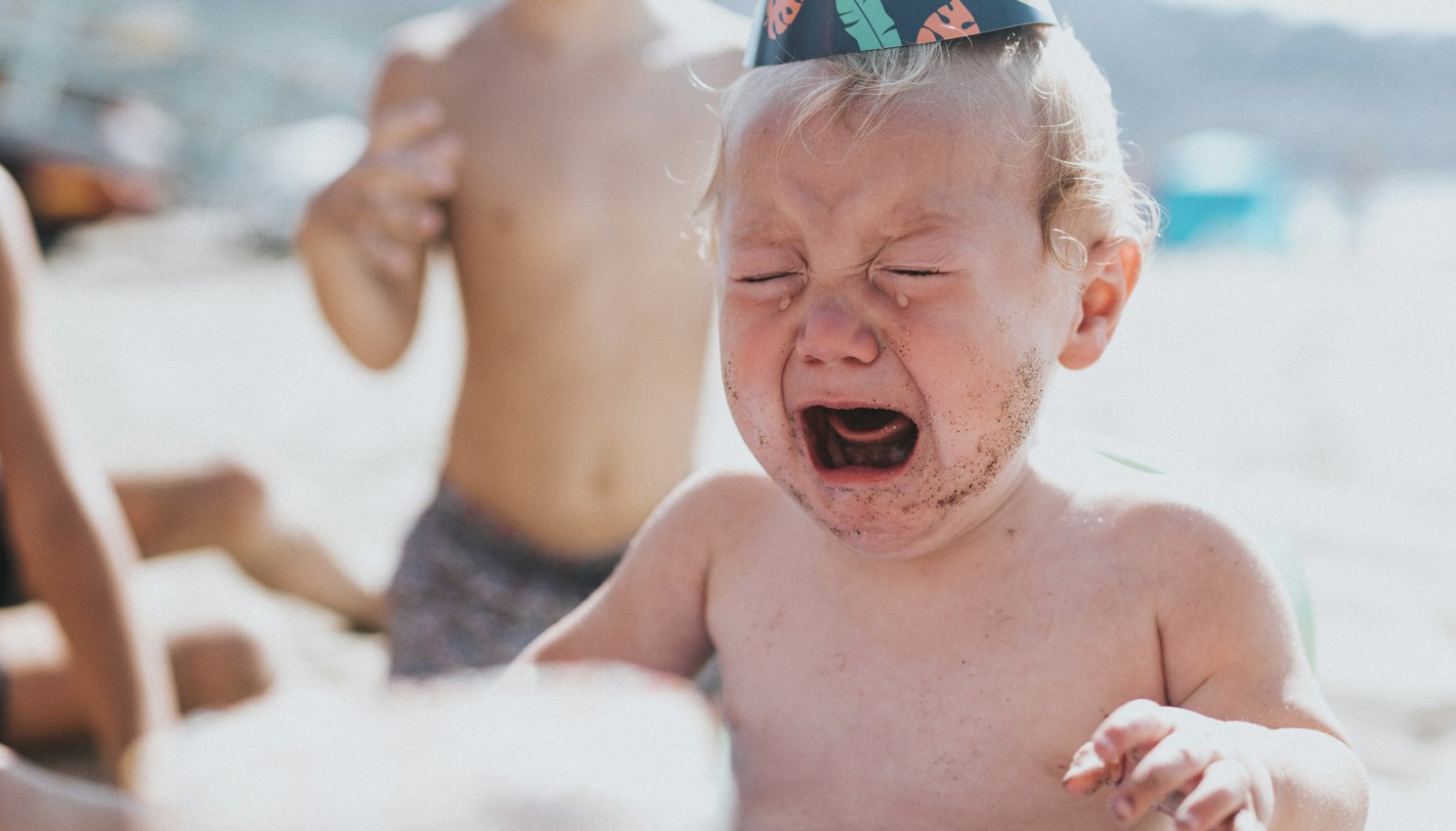 baby boy in blue and white floral shorts sitting on white and blue inflatable pool crying while wearing a party hat